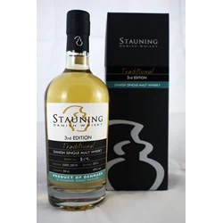 En flaske Stauning Danish Single Malt Whisky (3rd Edition) står ved siden af sin sorte og grå æske med guld- og blågrønne detaljer, begge med etiketten Traditional.